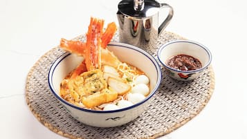 Un tazón de fideos con camarones, tofu y huevos cocidos, acompañado de un tazón pequeño de salsa y una olla de metal.