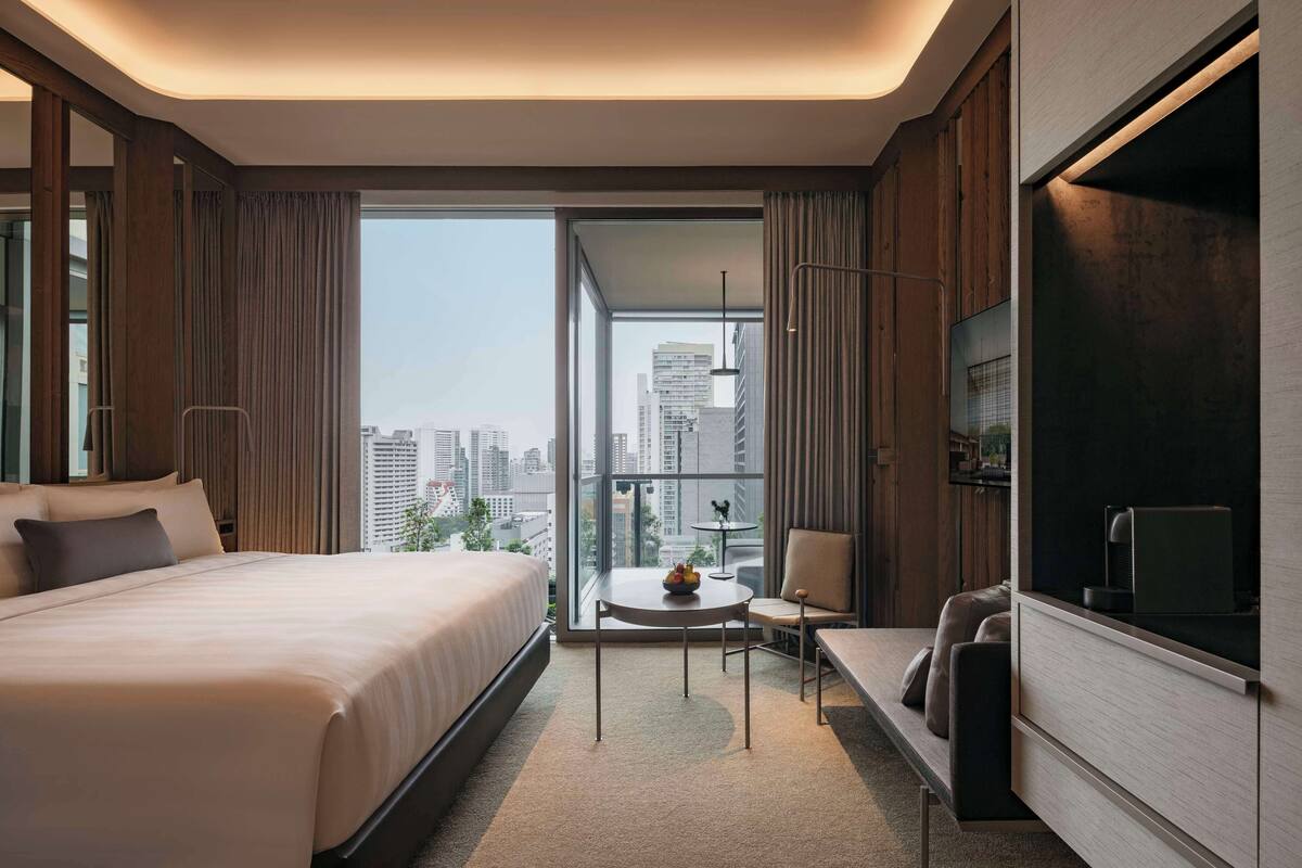 A modern hotel room with a large bed, a small round table with a fruit bowl, a chair, a TV, and a view of the city through large windows.