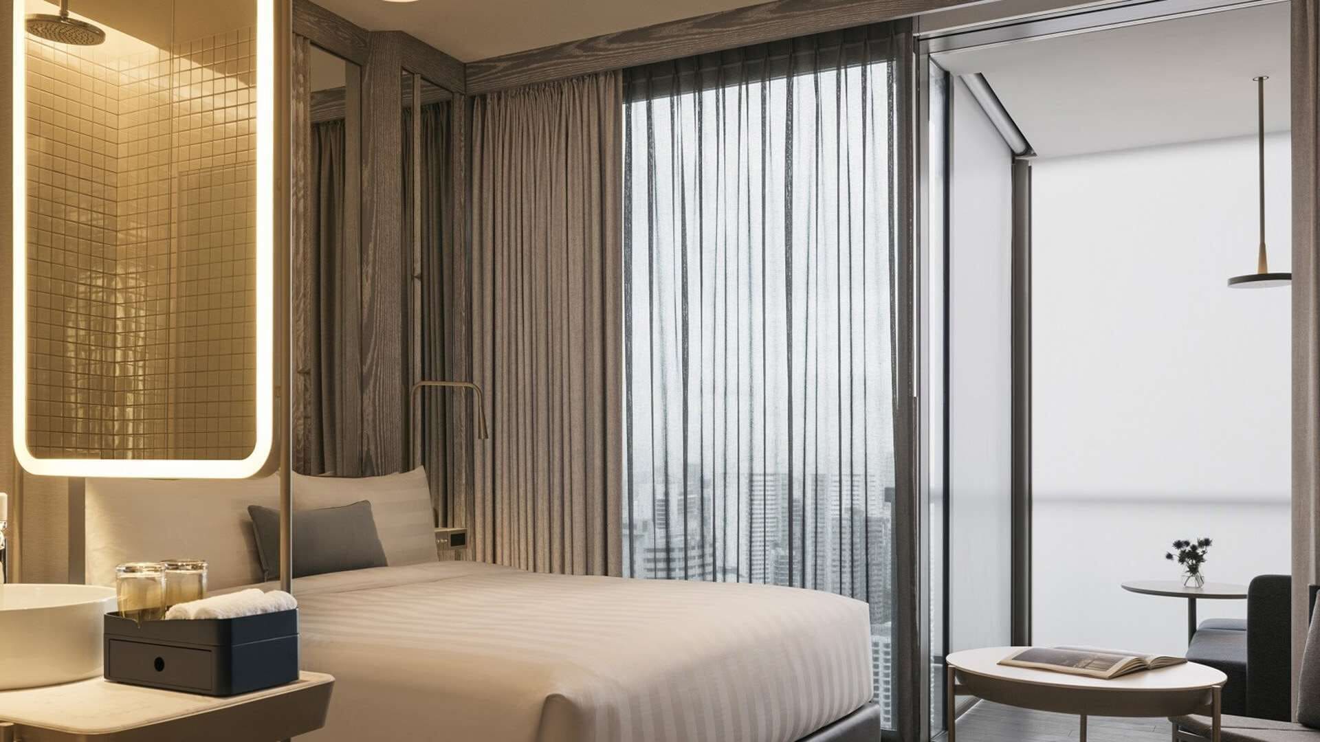A modern hotel room with a large bed, a bedside table with a lamp, a small table with a book, and a view of the city through the window.