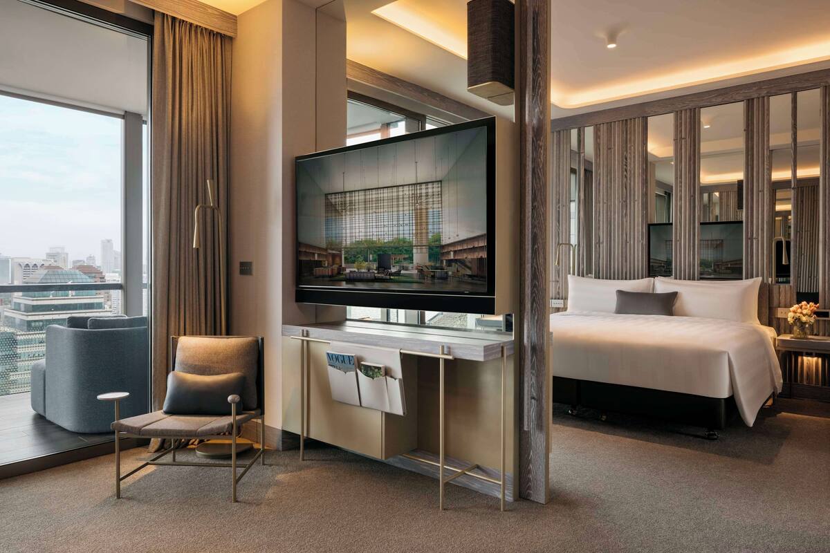 A modern hotel room with a large flat-screen TV, a comfortable chair, a neatly made bed, and a city view through the window.