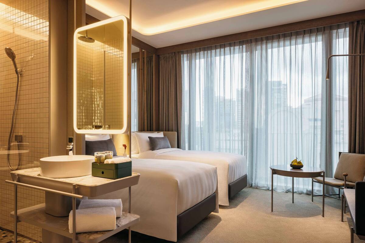 A hotel room with a large bed, a separate bathroom with a shower, a vanity with a sink, and a small table with a bowl of fruit.