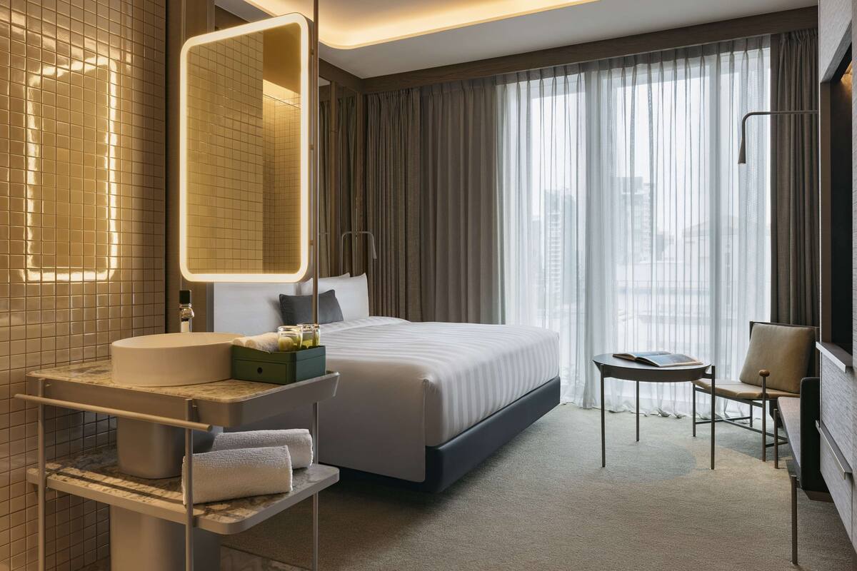 A modern hotel room with a large bed, a bedside table, a chair, a bathroom with a sink and mirror, and a view of the city through sheer curtains.