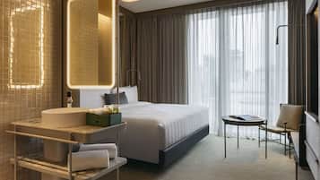 Habitación de hotel moderna con una cama grande, una mesita de noche, una silla, un baño con lavabo y espejo, y vistas de la ciudad a través de cortinas translúcidas.
