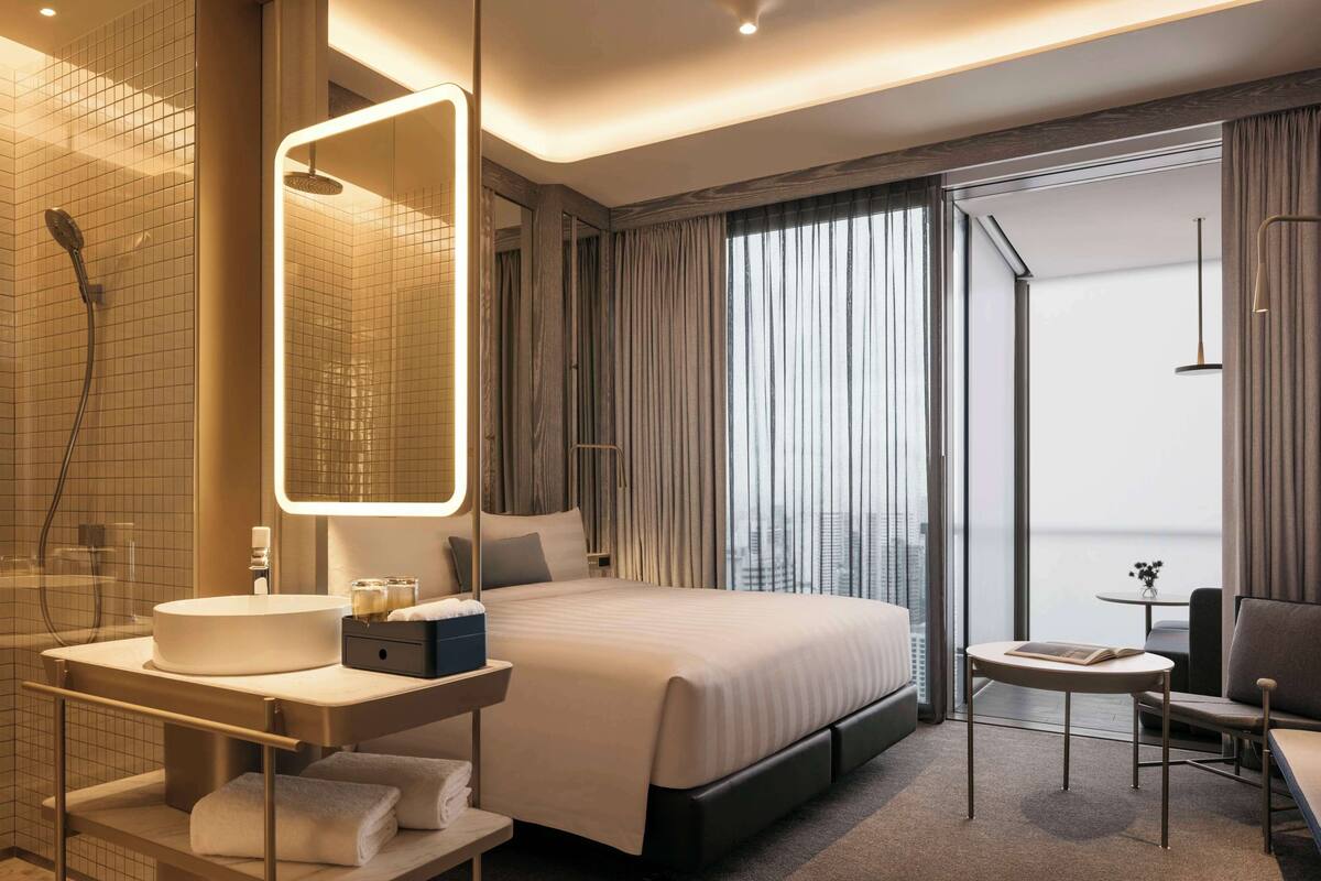 A modern hotel room with a large bed, a bedside table with a lamp, a bathroom with a shower and sink, and a view of the city through the window.
