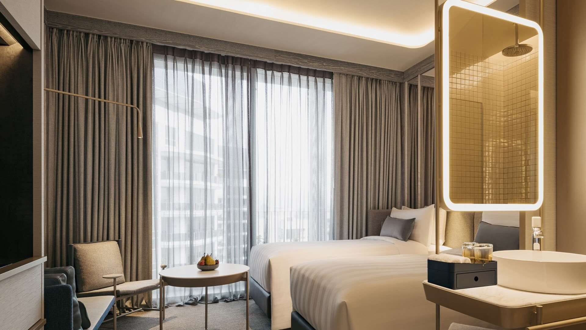 A hotel room with a large bed, a bedside table with a lamp, a chair, a small round table with a bowl of fruit, and a large mirror with a grid pattern.