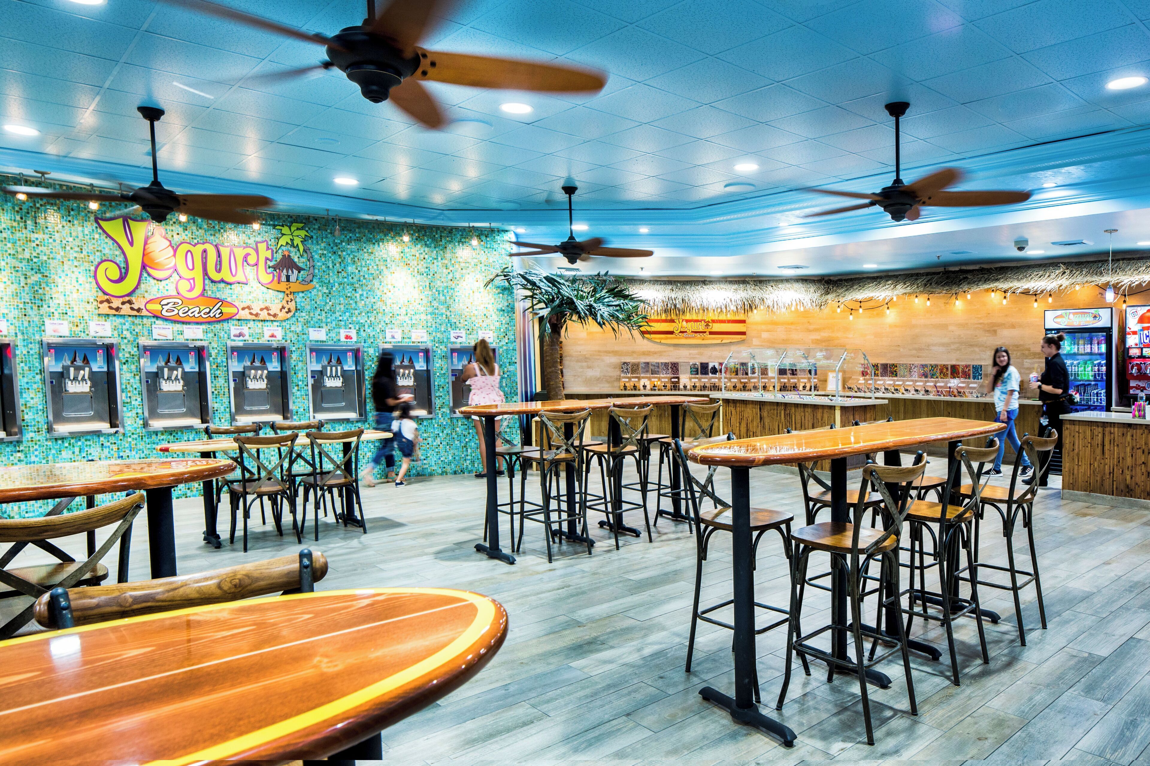 A restaurant interior with multiple tables and chairs, ceiling fans, and a colorful wall.