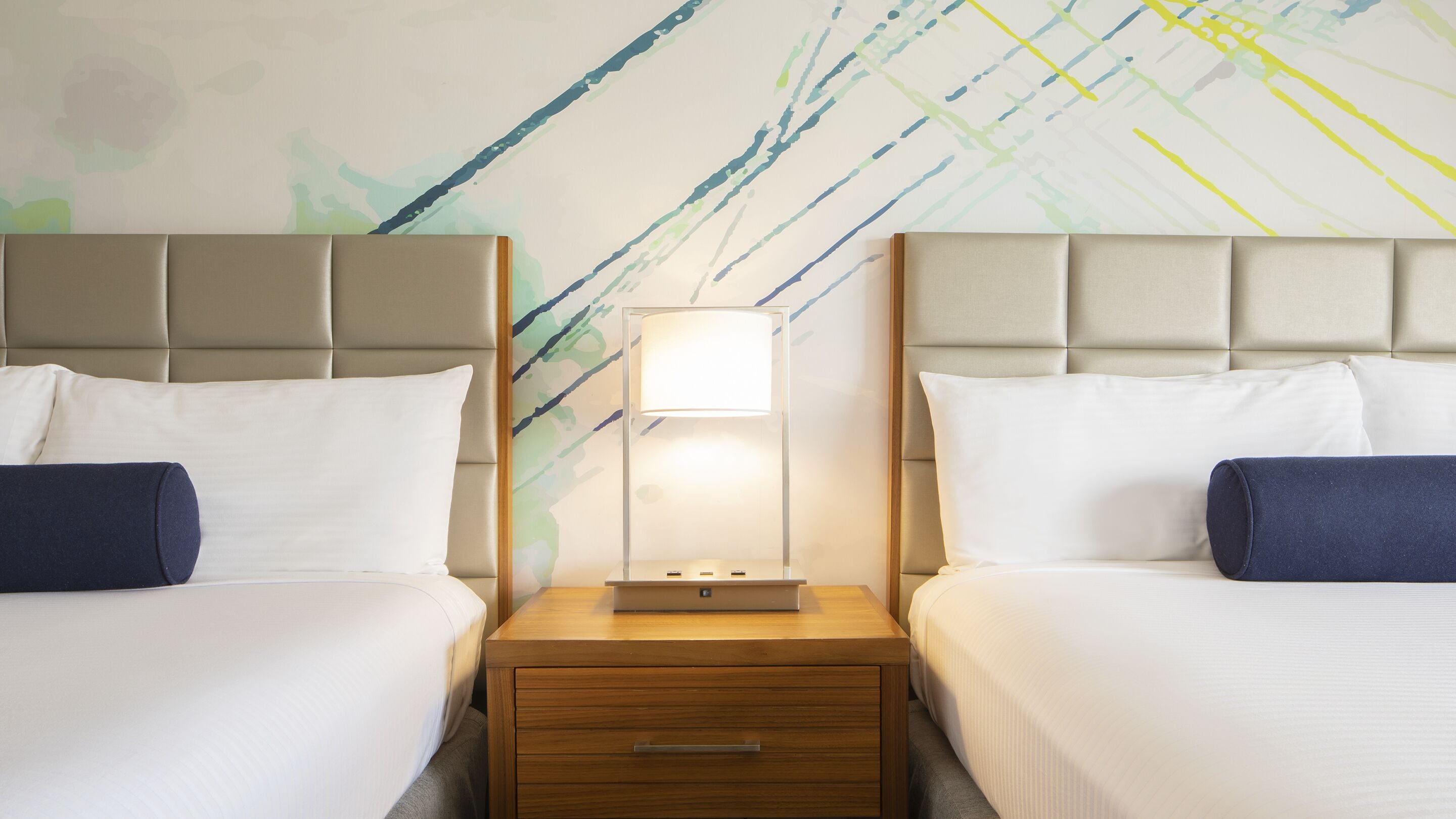 A hotel room with two beds, a nightstand with a lamp, and a wall with abstract art.
