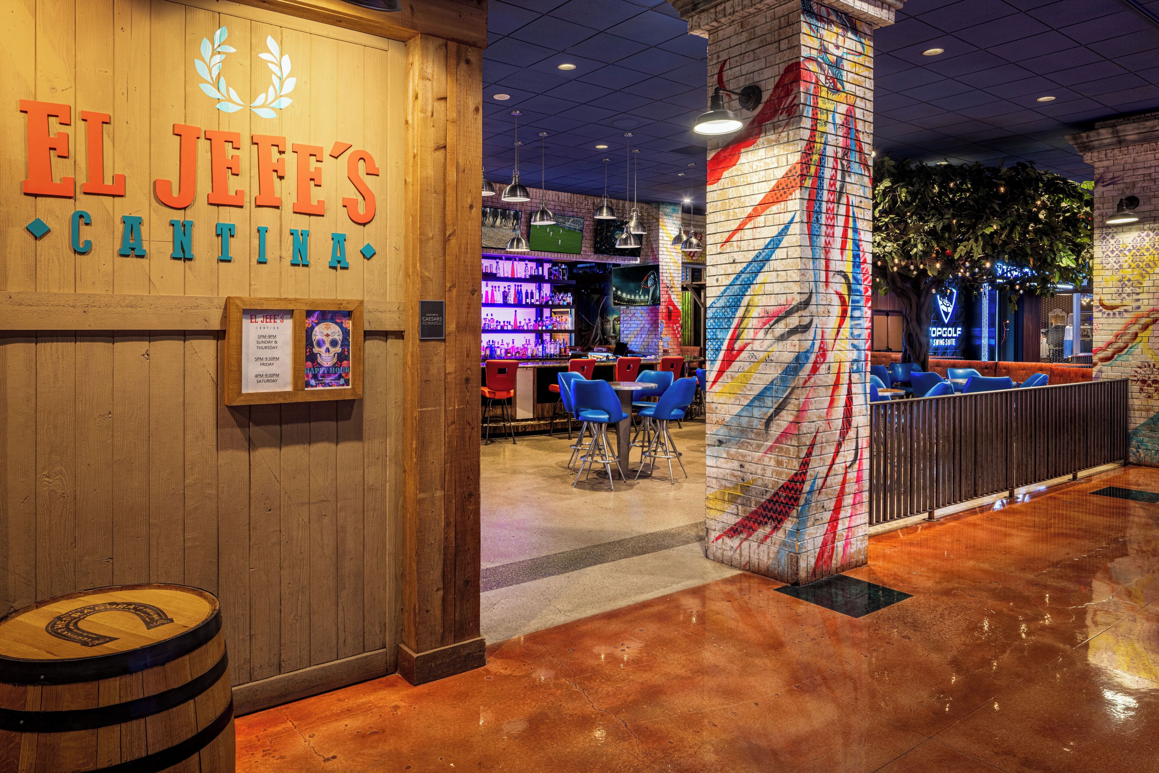 Una zona de bar con una pared de madera, un cartel que dice 'EL JEFF'S CANTINA' y un mural en la pared. Hay sillas azules y una barra con botellas.