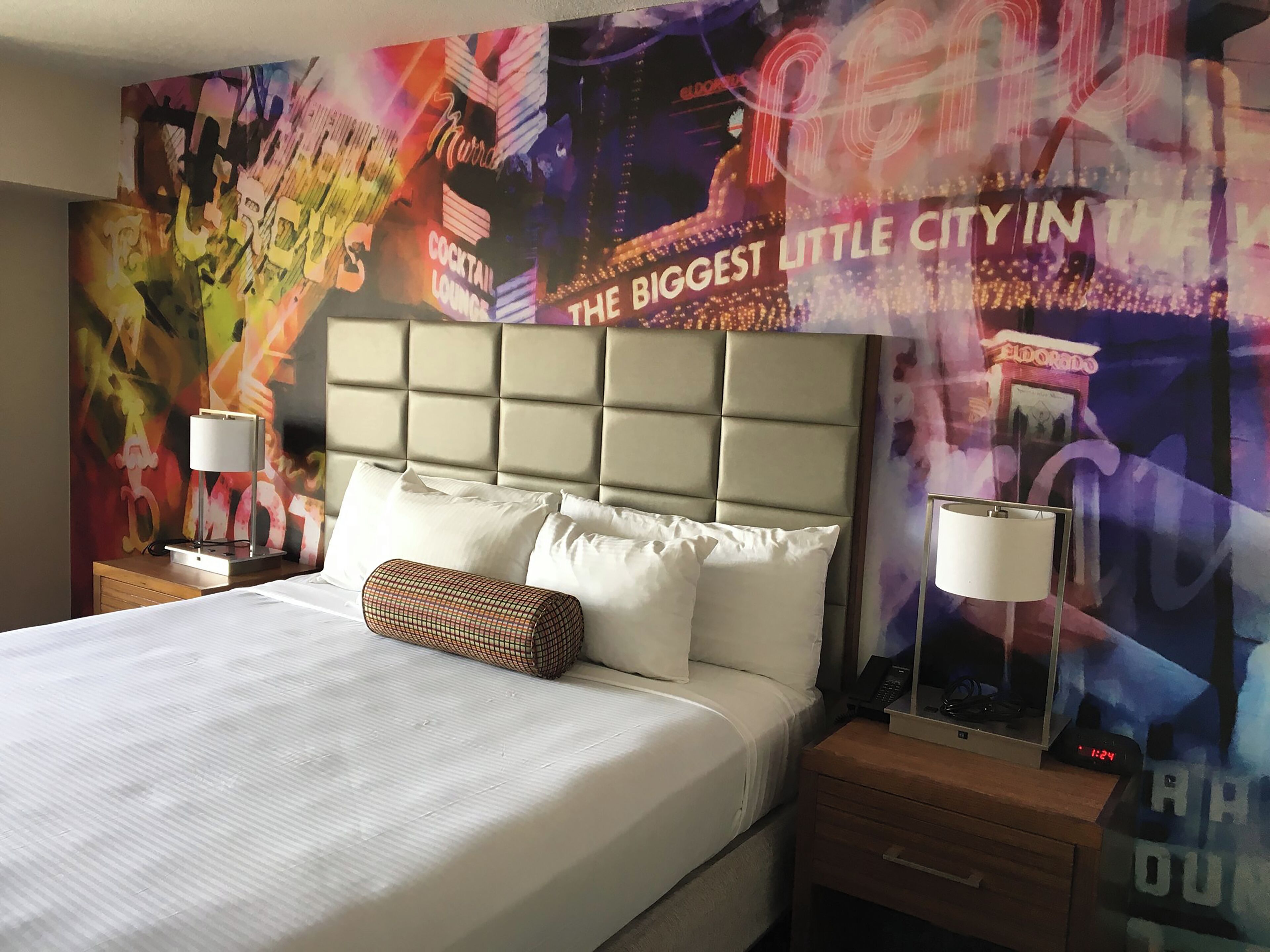 A hotel room with a large bed, two bedside tables, a headboard, and a vibrant mural on the wall.