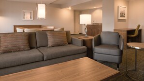 Ein modernes Hotelzimmer mit grauem Sofa, einem Holztisch, einem grauen Sessel und einem kleinen runden Tisch mit Lampe.