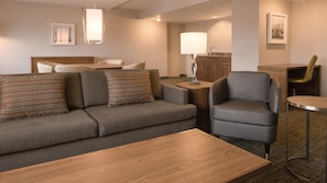 Een moderne hotelkamer met een grijze bank, een houten bijzettafeltje, een grijze fauteuil en een kleine ronde tafel met een lamp.