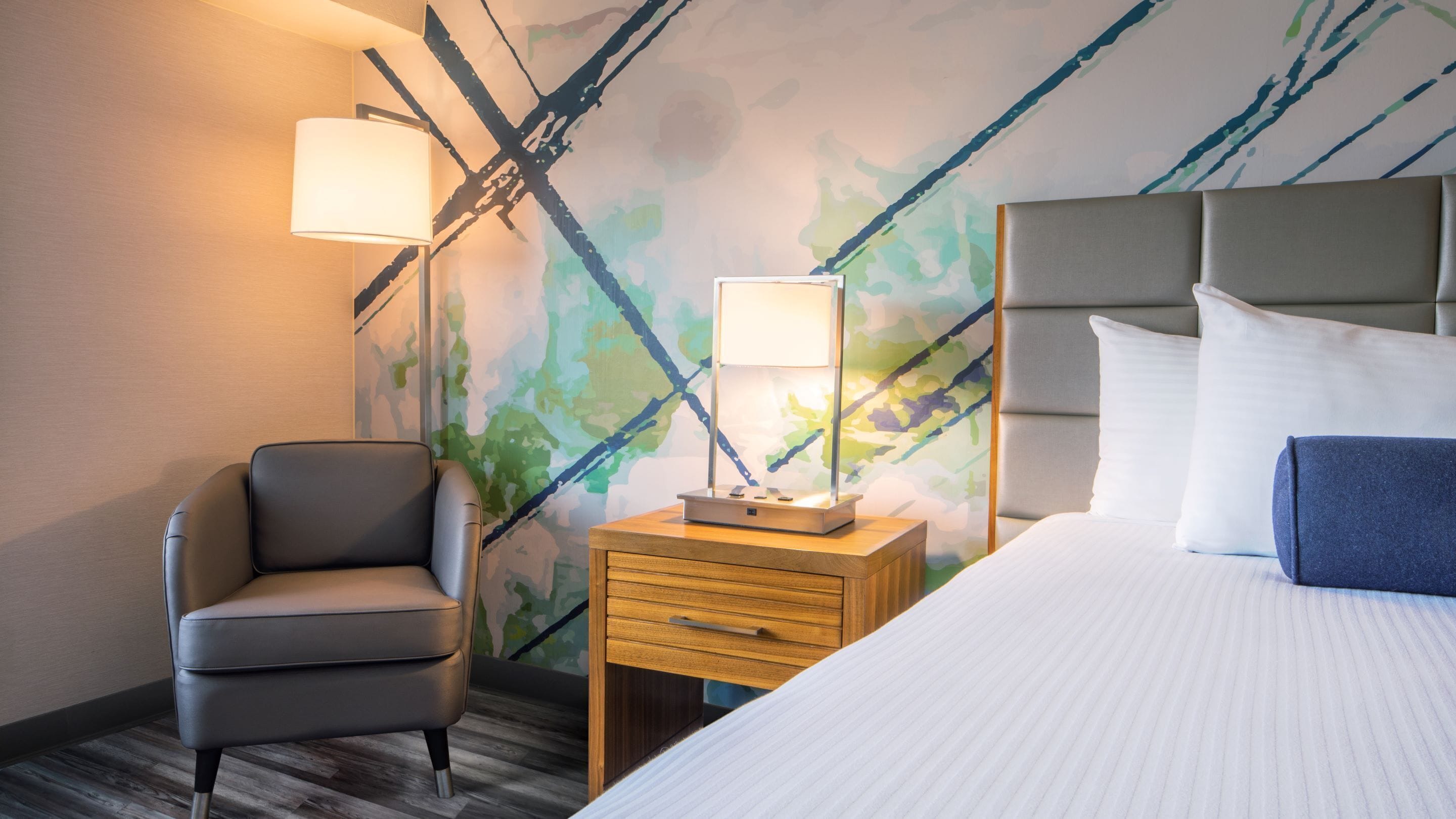 A hotel room with a bed, a chair, a nightstand with a lamp, and a wall with a camouflage pattern.