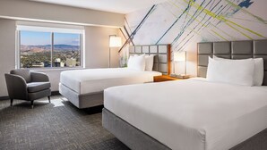 Ein Hotelzimmer mit zwei Betten, einem Sessel und einem großen Fenster mit Blick auf die Stadt.
