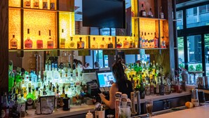 Una zona de bar con estantes llenos de botellas, un televisor montado en la pared y una barra con distintos objetos.