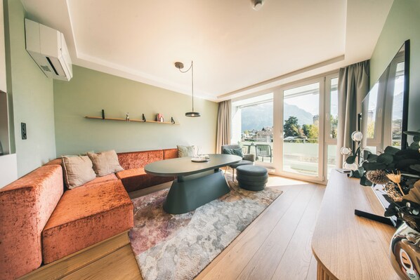 A modern living room with a sofa, a round table, and a large window offering a view of the outdoors. - HOP Apartments & Suites (Unterseen)