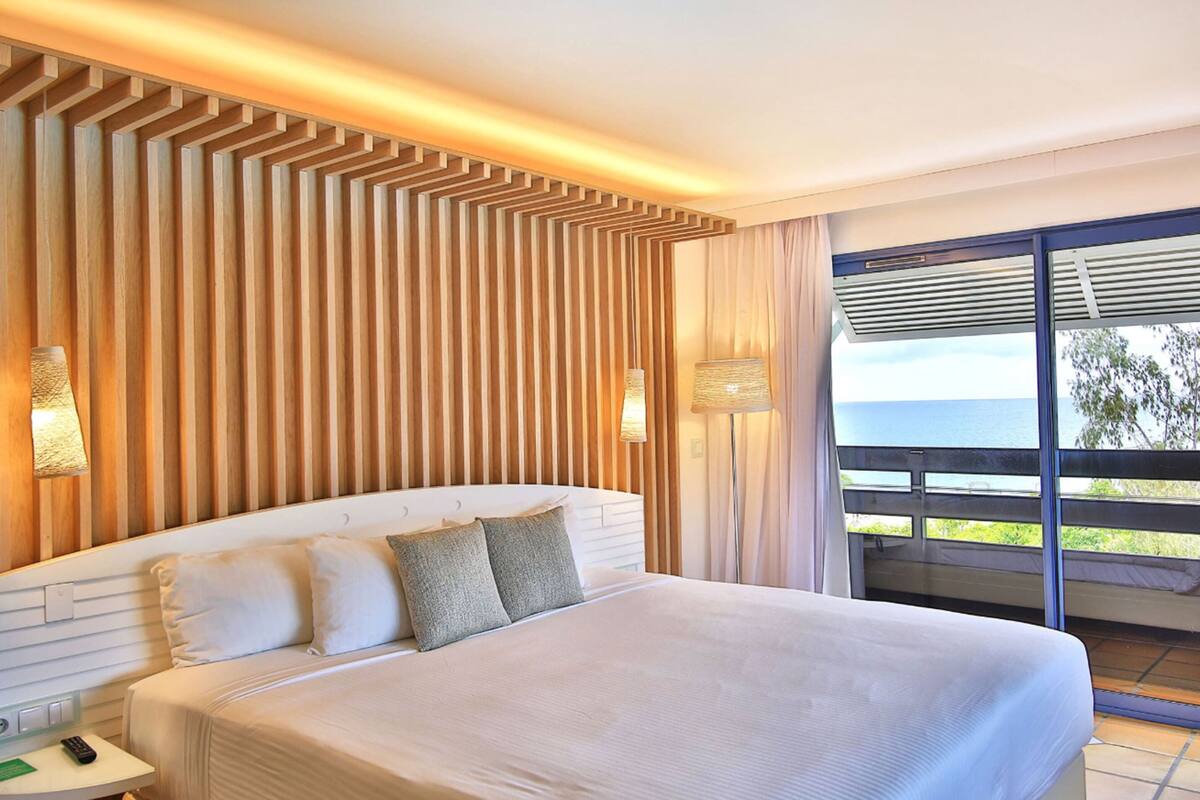 A modern hotel room with a large bed, wooden panel wall, and a balcony with a view of the sea.