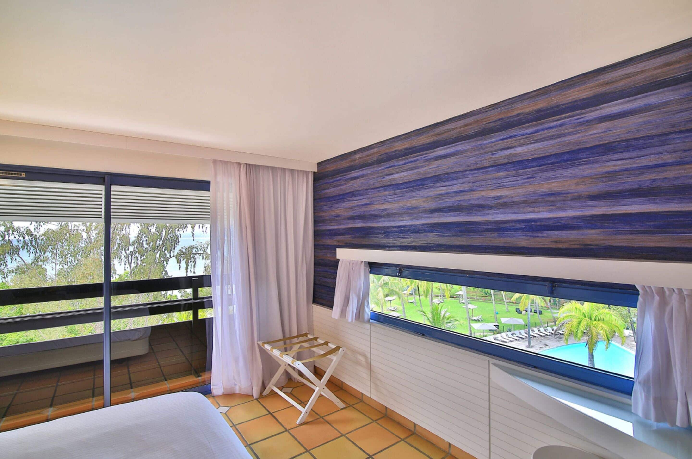 Quarto com varanda, cama, janela com vista para uma piscina e vegetação, e um varal de madeira.