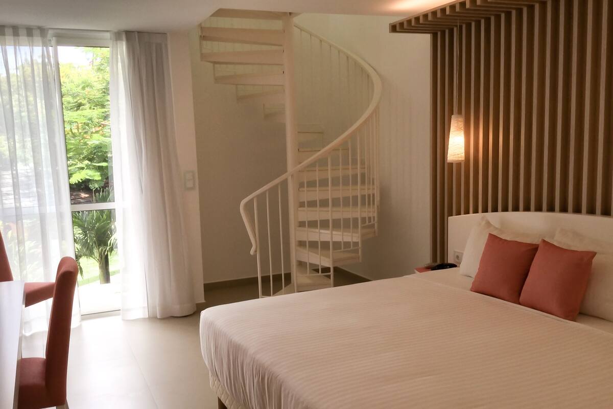 A bedroom with a large bed, a spiral staircase, and a view of greenery outside.