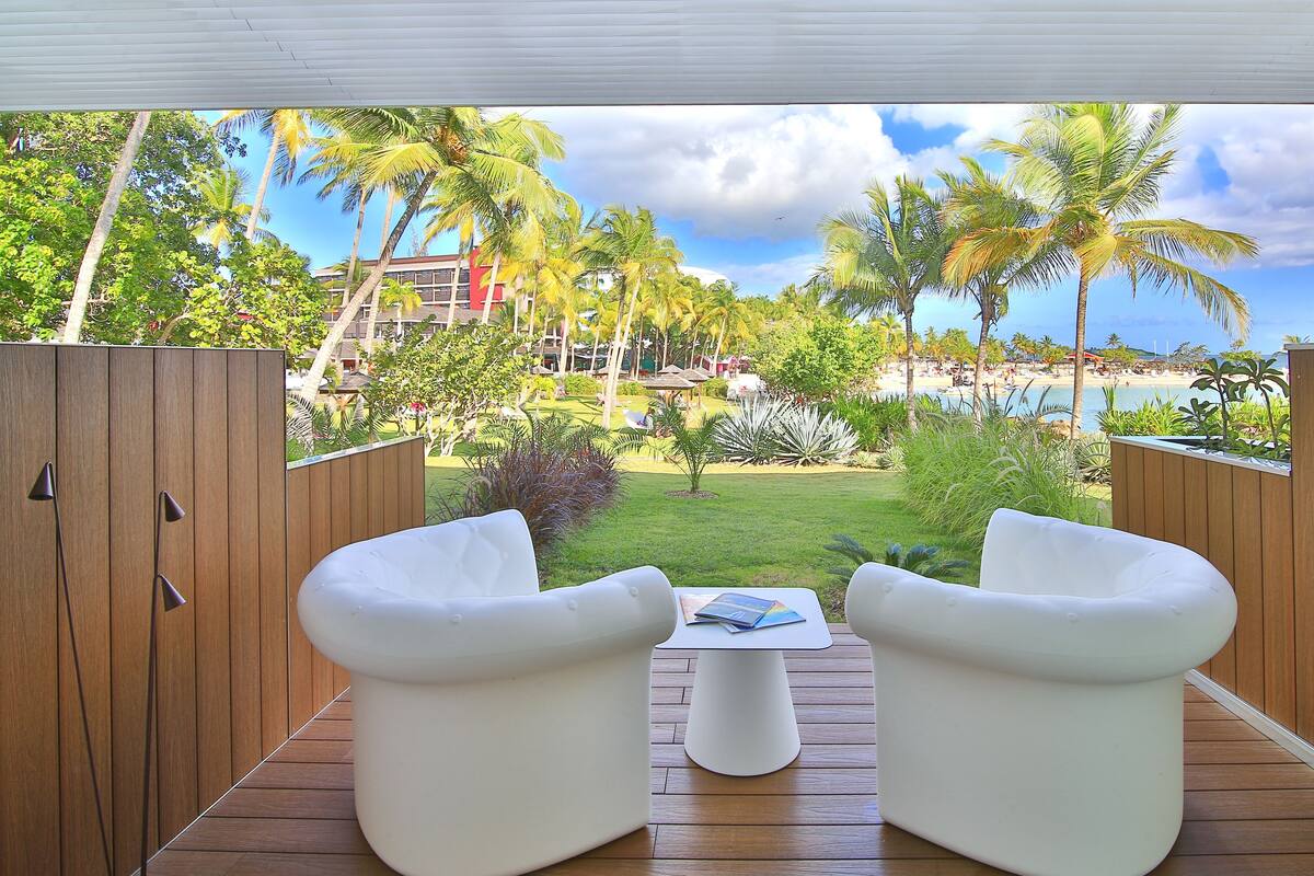 A deck with two white chairs and a small table, overlooking a beach and palm trees.
