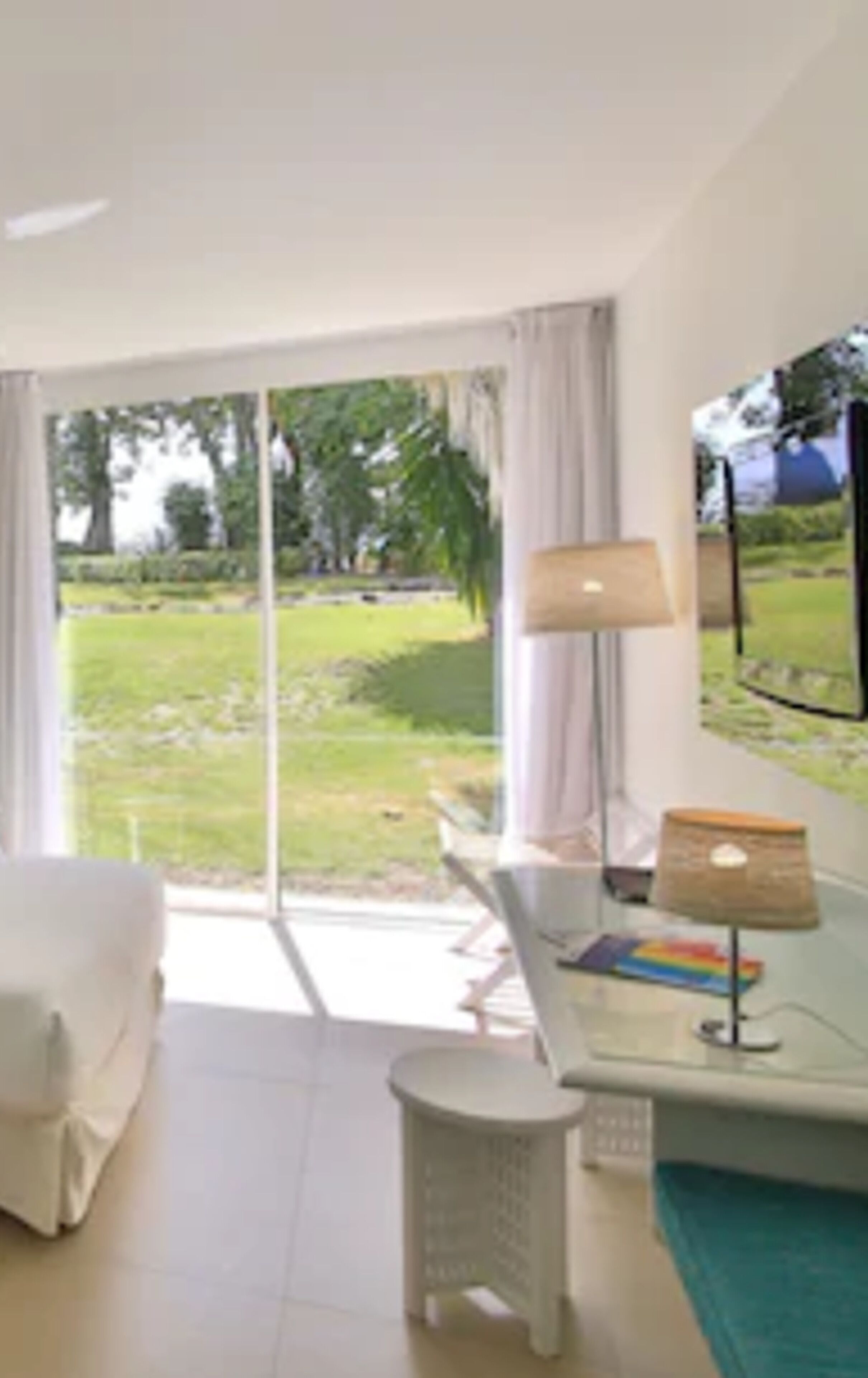 A modern room with a large sliding glass door leading to a garden. There is a white sofa, a small white table, and a lamp.