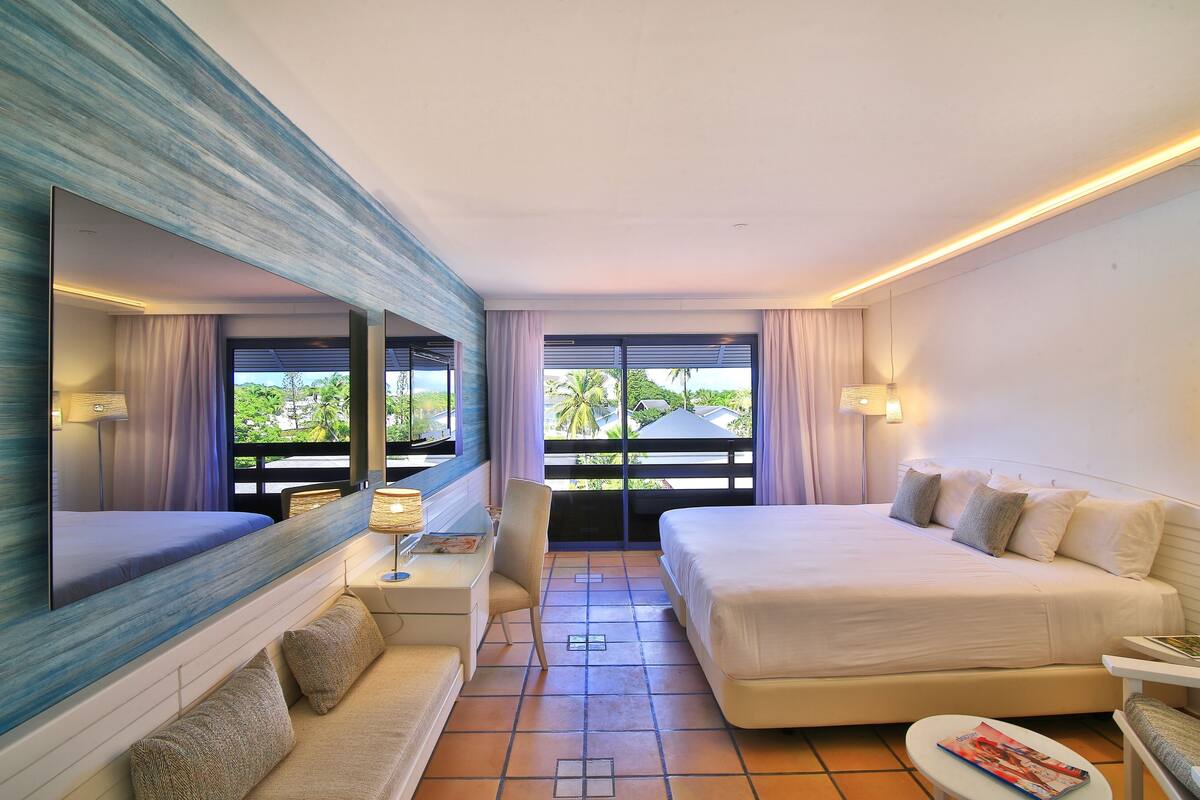 A hotel room with a large bed, a desk, a chair, a small table, and a large mirror. There are windows with curtains and a view of palm trees.