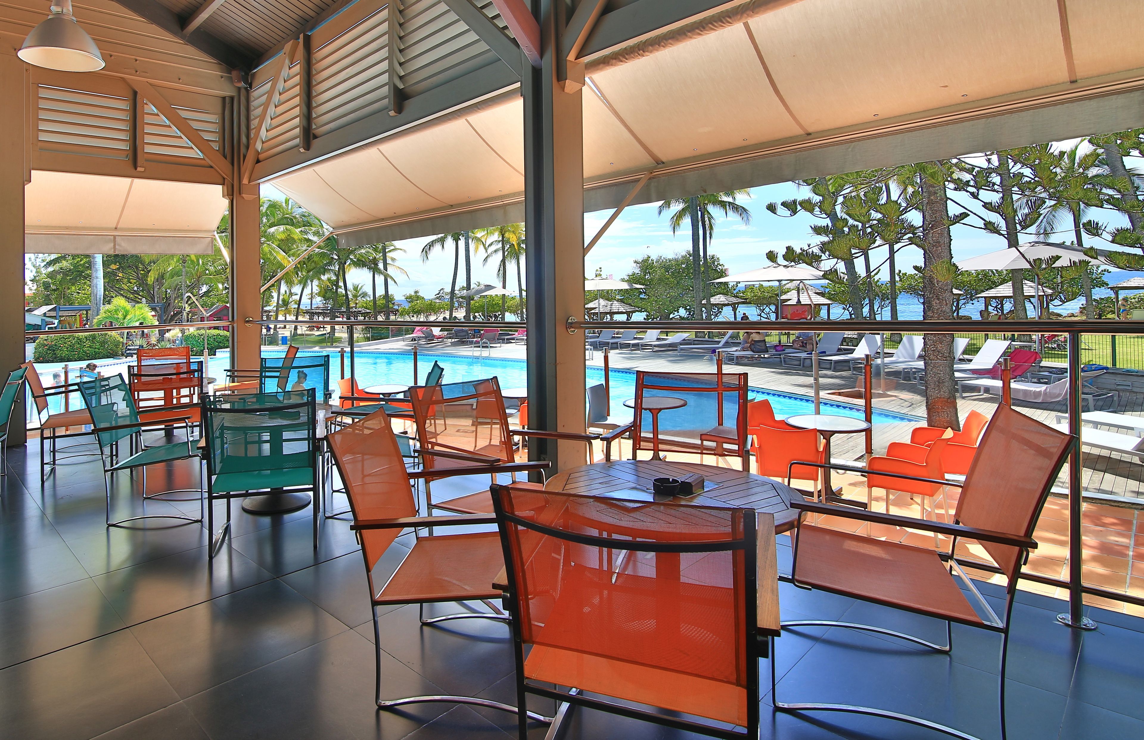 An outdoor seating area with tables and chairs, a pool, and palm trees in the background.