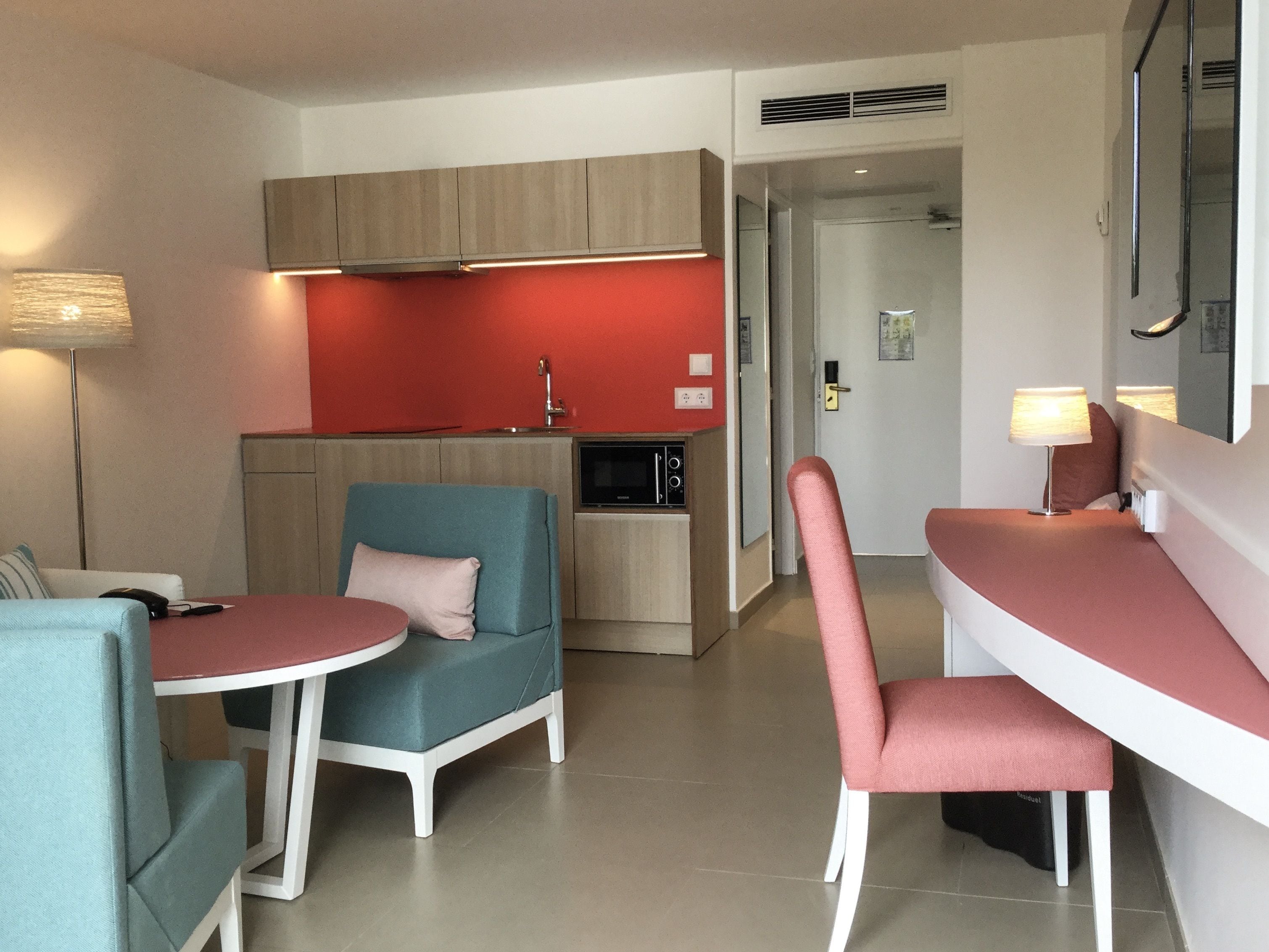 Una habitación de hotel compacta con kitchenette, un comedor con mesa y sillas, y un área de estar separada con sofá y sillón.