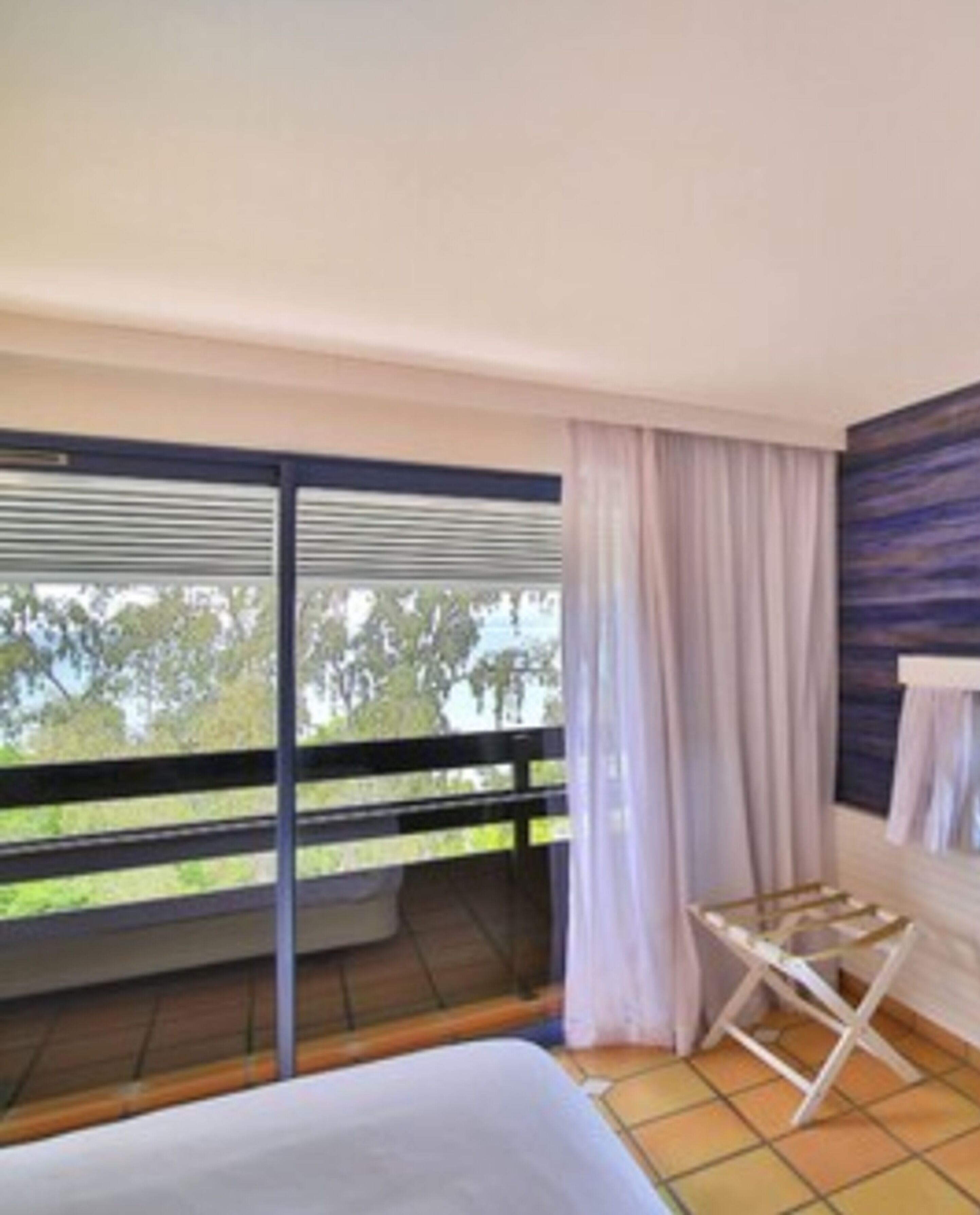 A hotel room with a balcony, a bed, a window with curtains, and a view of trees.