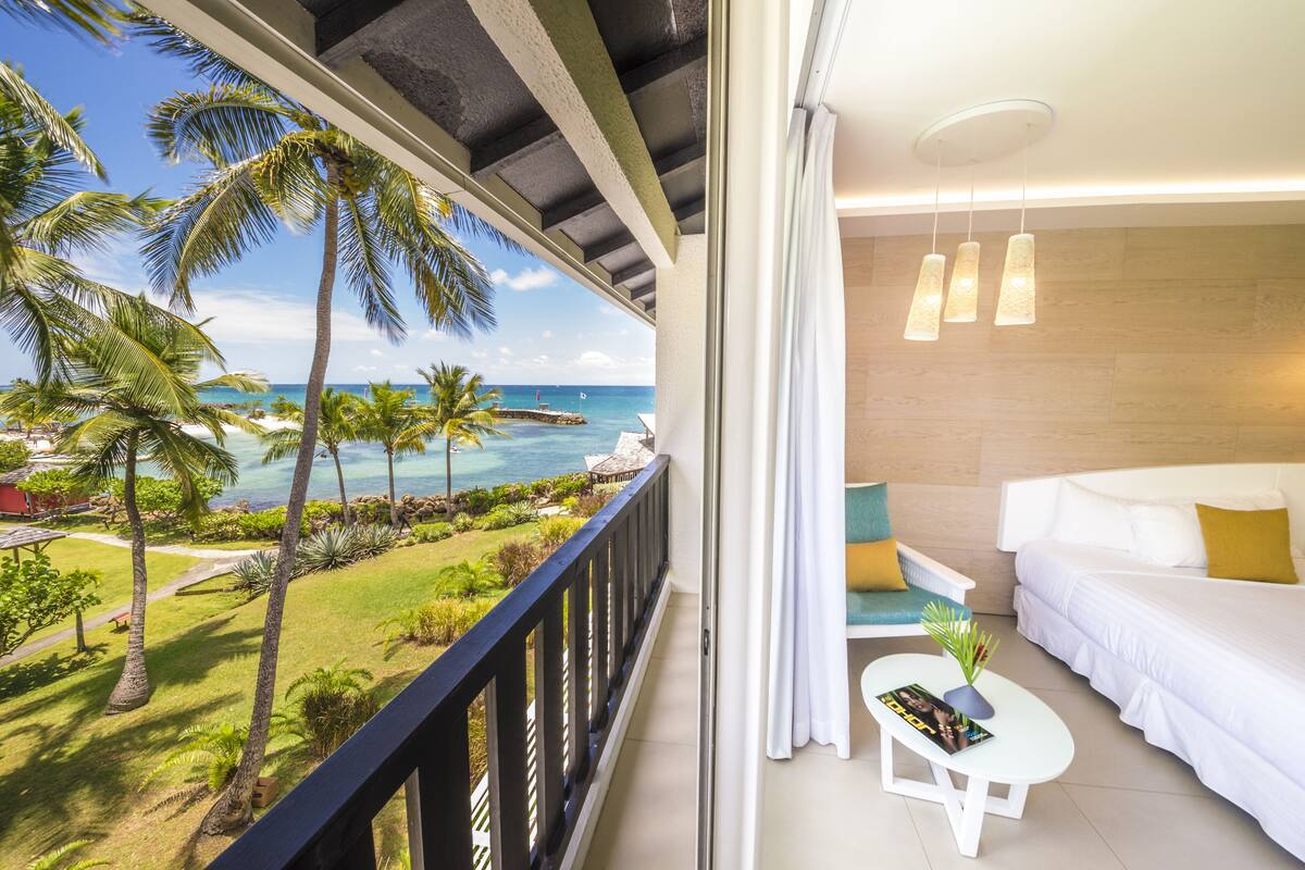 A balcony with a view of the ocean and palm trees, a white sofa, and a small table with a magazine.