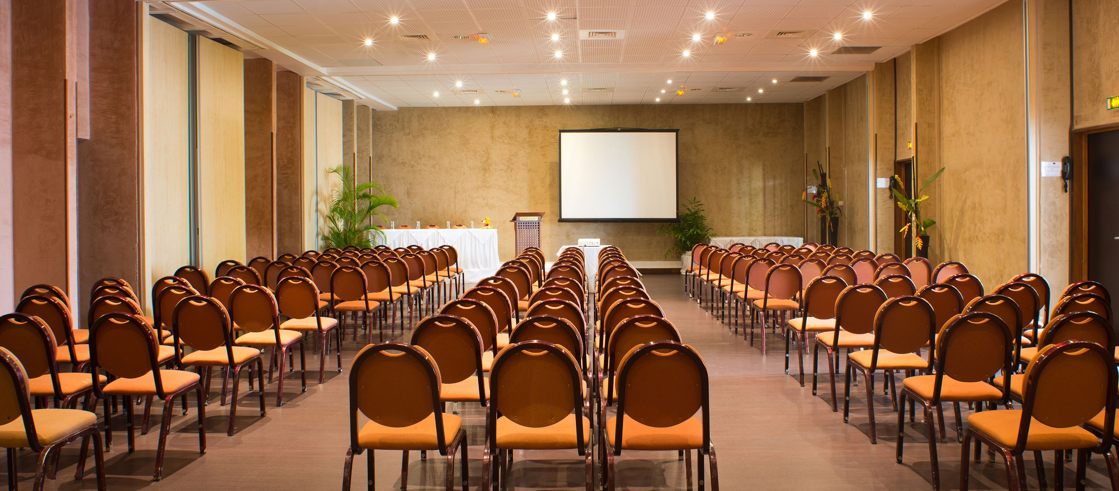 A conference room with rows of chairs facing a projection screen, a podium, and potted plants.