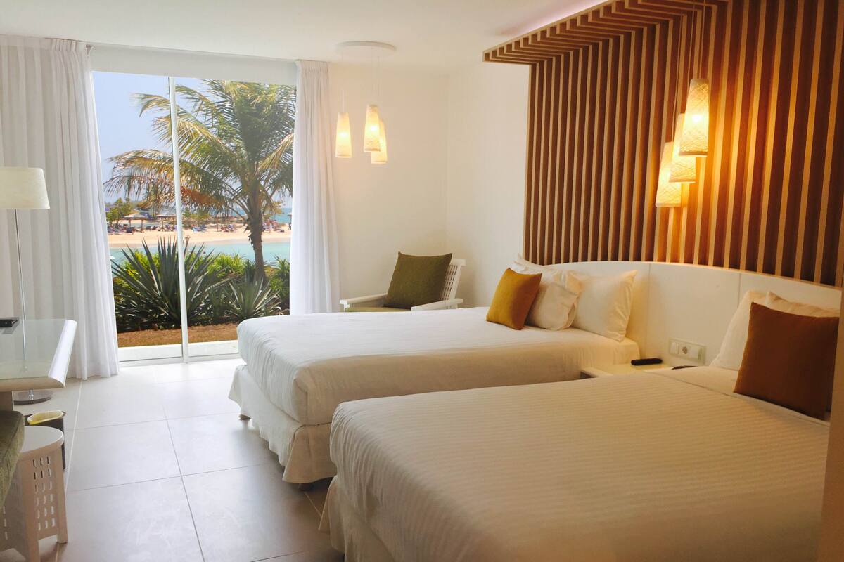 A hotel room with two beds, a desk, a chair, and a view of a beach and palm trees.