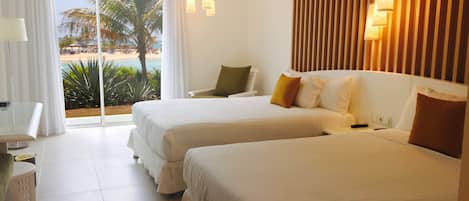 A hotel room with two beds, a desk, a chair, and a view of a beach and palm trees.