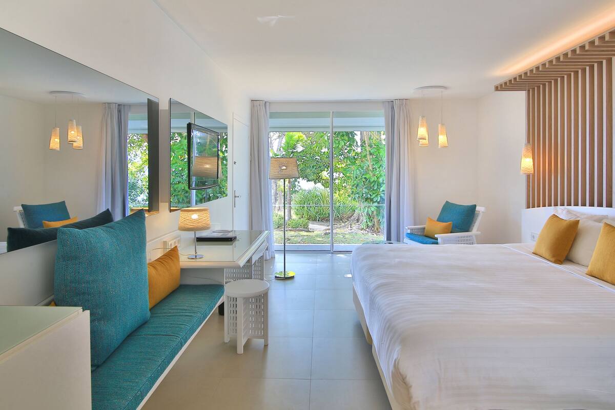 A modern hotel room with a large bed, a sofa, a small table, and a chair. There is a large window with a view of greenery outside.