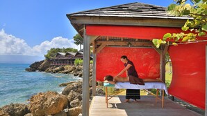 Sesión de masajes en una terraza de madera con vistas al océano, con un toldo rojo y vistas al mar y edificios cercanos.