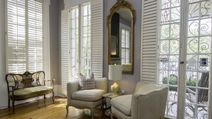 A room with a wooden floor, a green upholstered bench, two armchairs, a small table with a lamp, and a large mirror on the wall. - Rathbone Mansions (New Orleans)