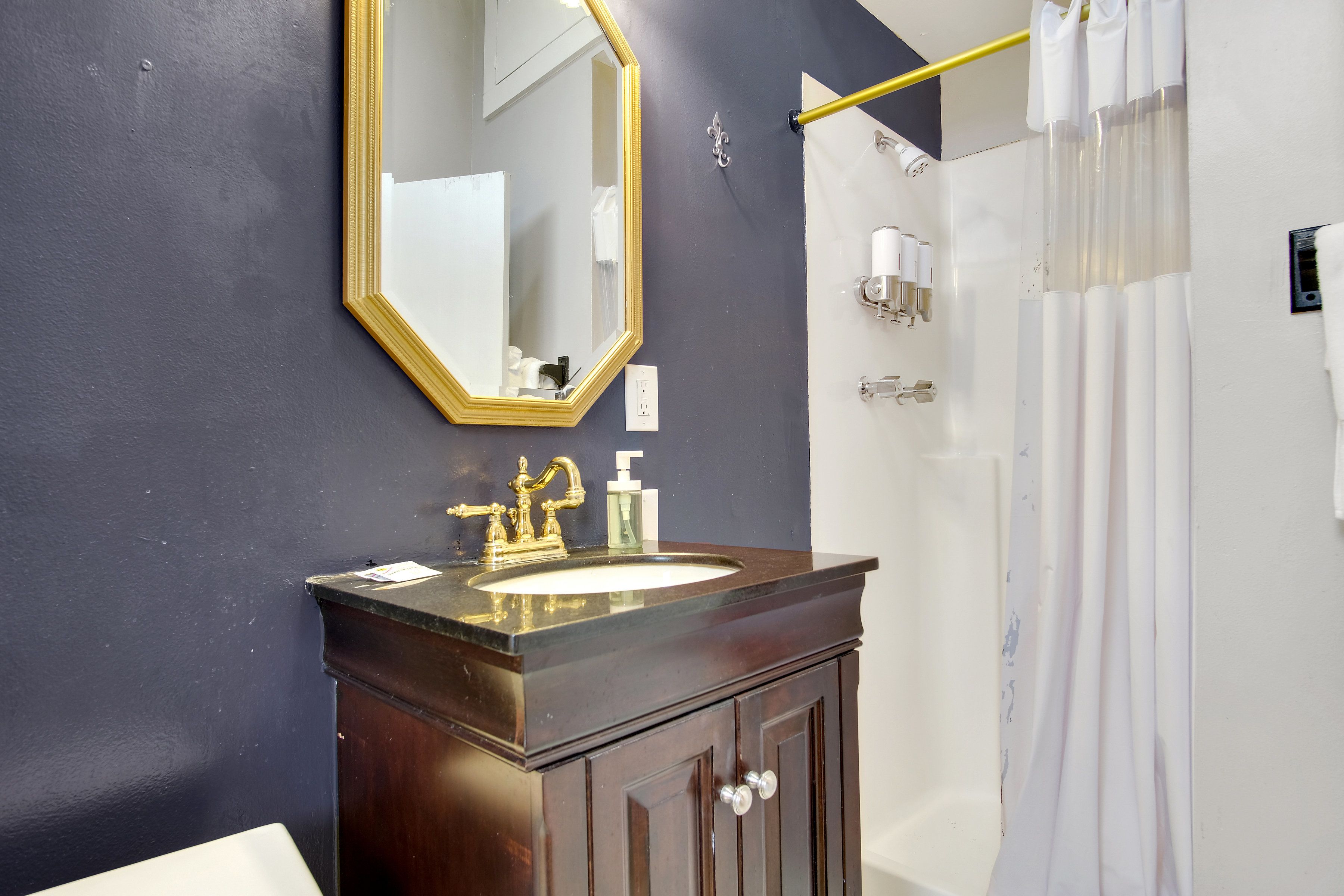Un baño con una pared oscura, un espejo con marco dorado, un mueble de tocador de madera oscura, un lavabo blanco y una ducha con cortina blanca.