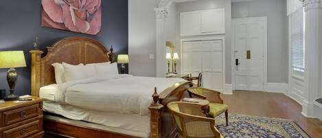 A bedroom with a large bed, wooden headboard, and a decorative mirror above it. There is a nightstand with a lamp and a small table with a chair.