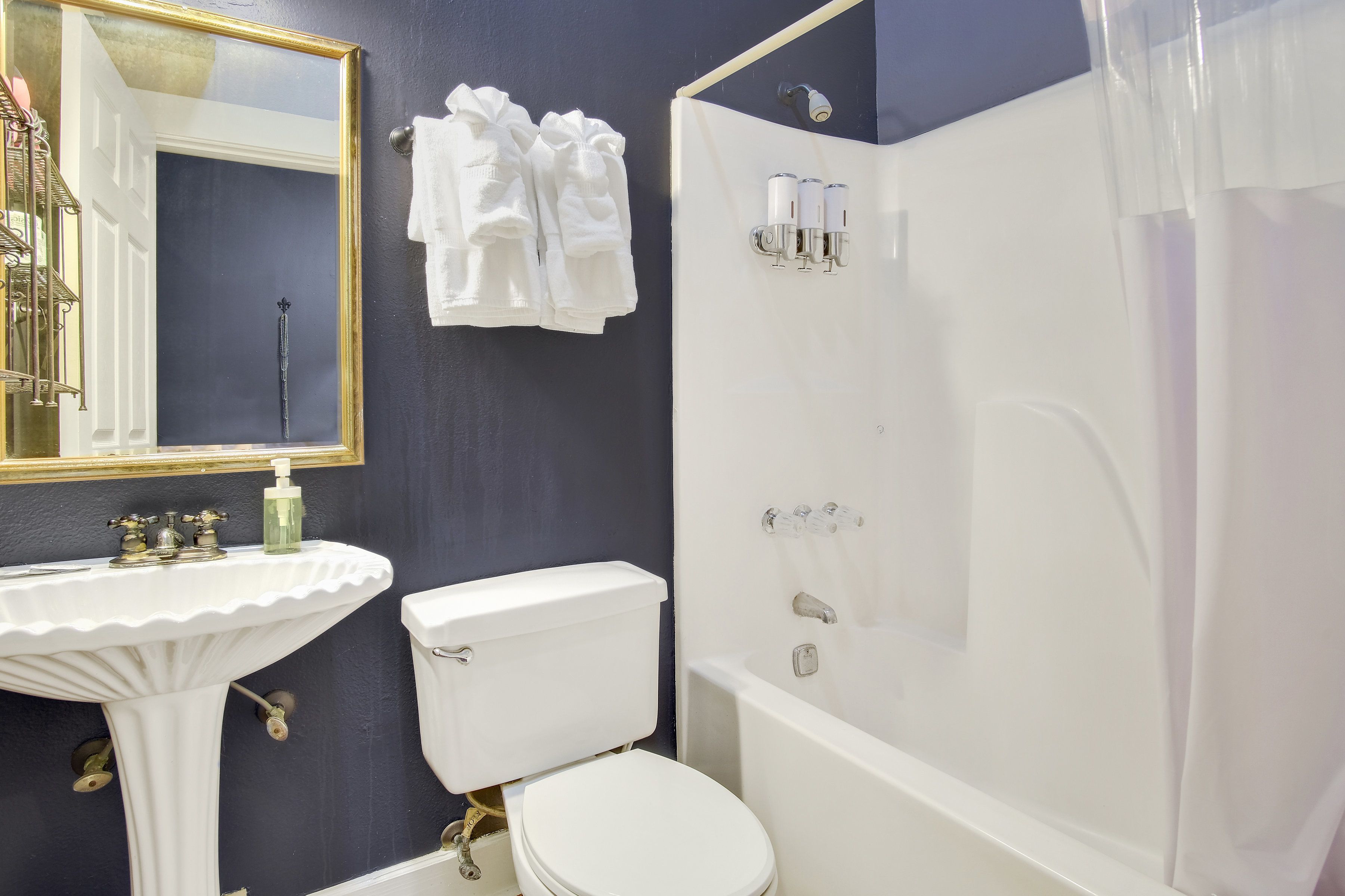 Un baño con lavamanos, inodoro y bañera blancos. Hay un espejo con marco dorado y toallas colgadas en la pared.