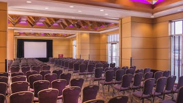 Un salón de conferencias con filas de sillas moradas, un escenario y una pantalla grande.