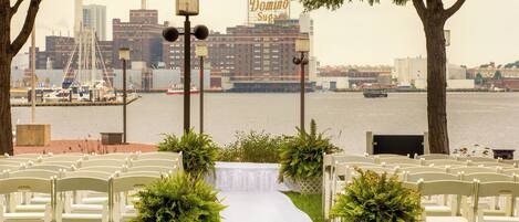 A wedding aisle with white chairs and a white carpet, leading to a cityscape with a Domino Sugar sign in the background.