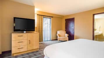 Habitación de hotel con una cama, un televisor sobre un mueble de madera, una silla, una puerta y una ventana con cortinas.