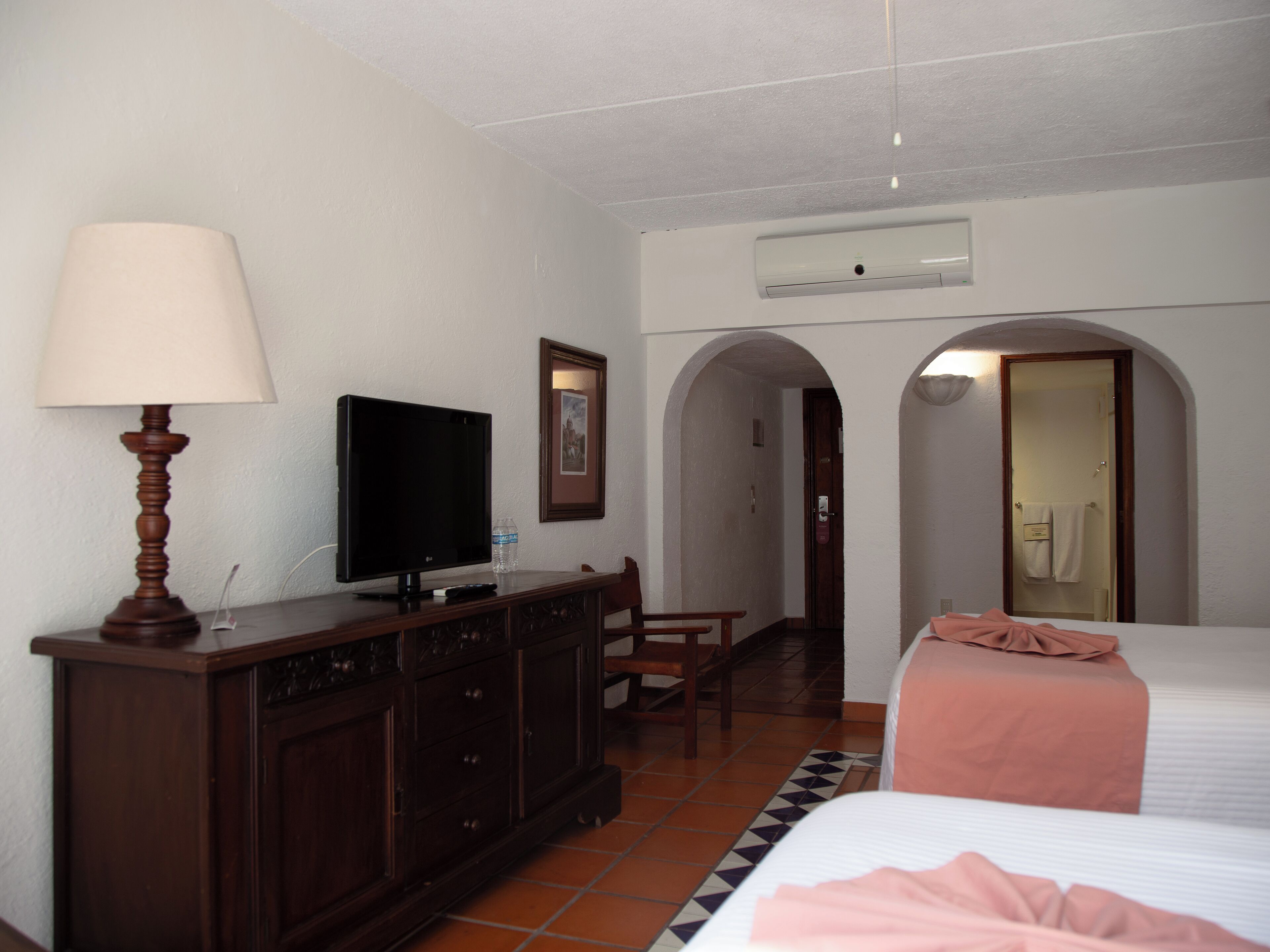 A hotel room with a bed, a TV on a wooden dresser, a lamp, and a bathroom visible through an archway.