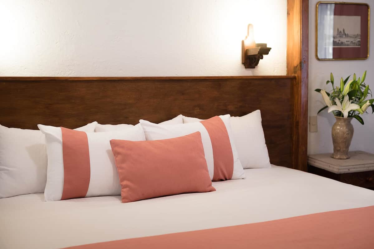 Una cama bien tendida con almohadas blancas y melocotón, un cabecero de madera, una mesita de noche con un florero y un cuadro en la pared.
