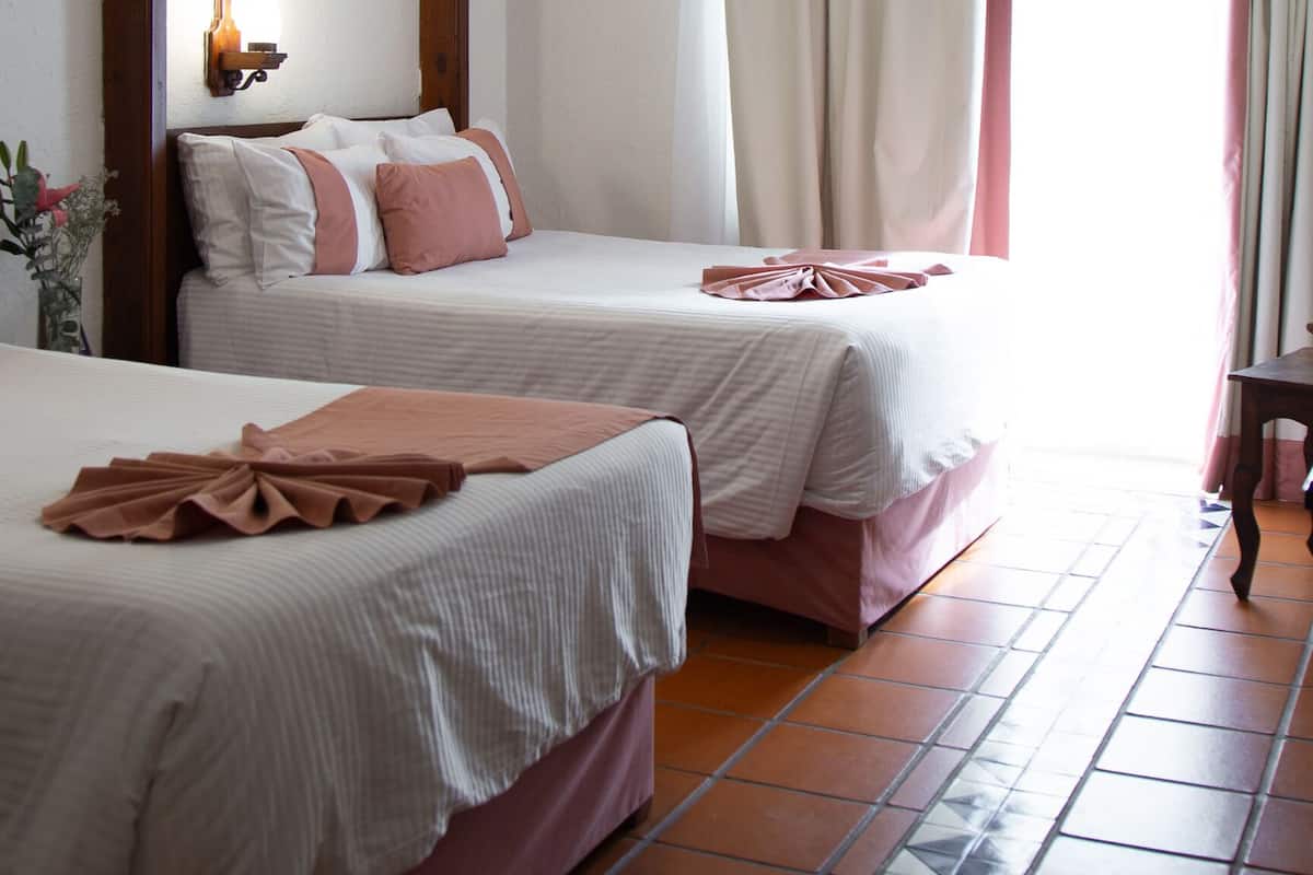 Habitación de hotel con dos camas, un ventilador de techo, una ventana con cortinas y una lámpara fijada a la pared.