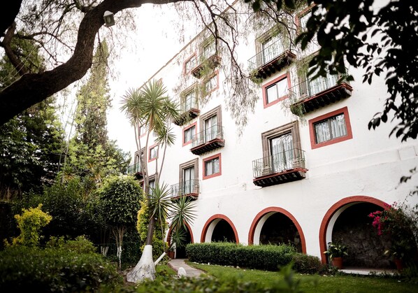 A white building with red accents, multiple balconies, and a lush garden with various plants and a pathway. - Hotel Misión Guanajuato (Guanajuato)