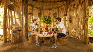 Deux personnes sont assises à une table ronde, sous un toit de chaume et entre des murs en bambou, dans un décor naturel agrémenté de meubles simples.