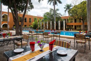 An outdoor dining area with tables set for two, chairs, and a pool in the background.