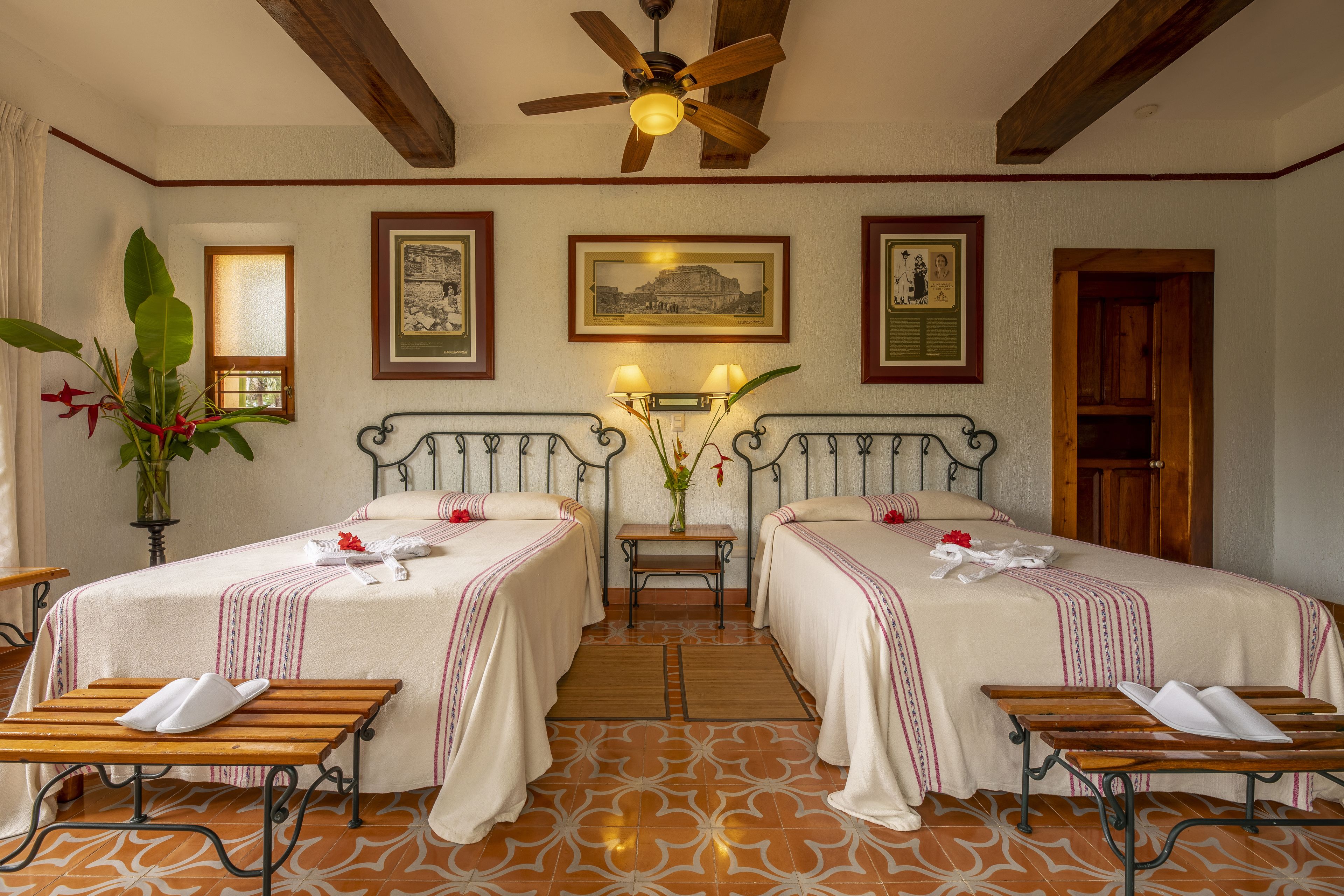 A hotel room with two beds, a ceiling fan, a small table with a vase of flowers, and a wooden bench.