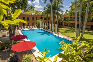 A swimming pool with lounge chairs and umbrellas, surrounded by palm trees and a building with arched windows. - Hacienda Uxmal Plantation & Museum (Santa Elena)