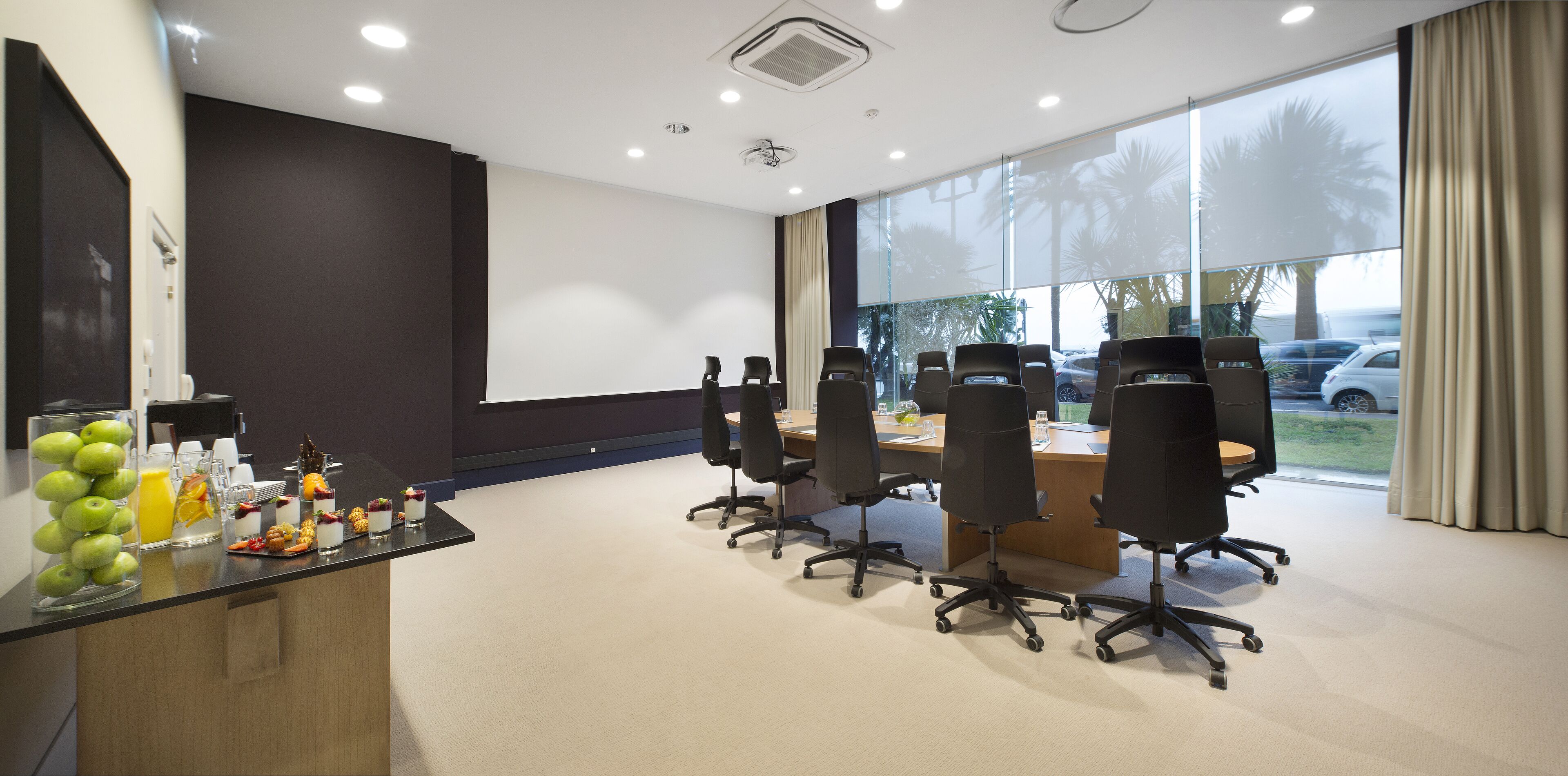 A conference room with a long table, multiple chairs, a glass display case with fruits, and a projector screen.